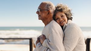 Romantic senior woman smiling at the camera while embracing her husband by the ocean. Affectionate elderly couple enjoying spending some quality time together after retirement.